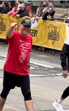 John Cressler walking in the Pittsburgh Marathon in a red shirt and black hat pointing at the camera while smiling.