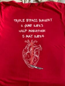 Front of John Cressler's red shirt he wore in Pittsburgh Marathon. Text saying 'triple bypass surgery 2 june 2023, Half marathon 5 May 2024, above a line illustration of a heart with a scalpel at the bottom of the heart.